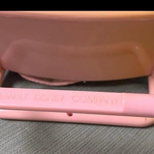 May include: A light pink plastic toy with the text "WALT DISNEY COMPANY" embossed on the front. The toy has a rounded shape and appears to be a part of a larger set. The background is a muted green fabric.