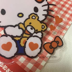 Pode incluir: Estojo de pl&aacute;stico transparente com Hello Kitty e um ursinho de pel&uacute;cia. O design inclui um grande la&ccedil;o, cora&ccedil;&otilde;es e o texto "'76, '17 SANRIO". O estojo est&aacute; em um fundo xadrez vermelho e branco.
