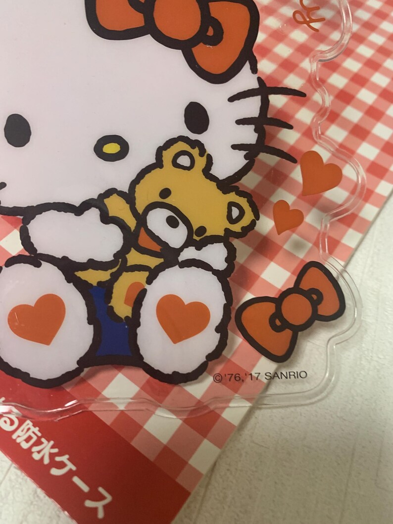 Pode incluir: Estojo de pl&aacute;stico transparente com Hello Kitty e um ursinho de pel&uacute;cia. O design inclui um grande la&ccedil;o, cora&ccedil;&otilde;es e o texto "'76, '17 SANRIO". O estojo est&aacute; em um fundo xadrez vermelho e branco.