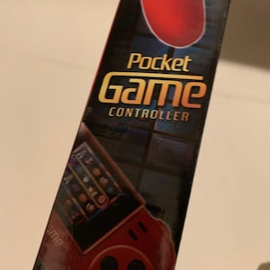 Pode incluir: Uma caixa preta e vermelha do Pocket Game Controller. A caixa apresenta um gr&aacute;fico de controle de jogo vermelho na parte superior e um gr&aacute;fico menor de um console de jogo. As palavras "Pocket Game Controller" s&atilde;o em ouro e branco.