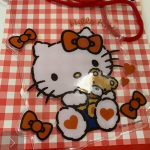 Pode incluir: Capa de telefone transparente com Hello Kitty segurando um ursinho de pel&uacute;cia, com la&ccedil;os e cora&ccedil;&otilde;es laranja em um fundo xadrez vermelho e branco. A capa tem um cord&atilde;o vermelho e o texto "Hello Kitty".