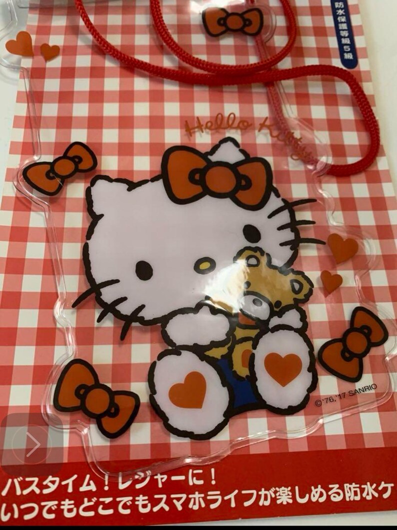 Pode incluir: Capa de telefone transparente com Hello Kitty segurando um ursinho de pel&uacute;cia, com la&ccedil;os e cora&ccedil;&otilde;es laranja em um fundo xadrez vermelho e branco. A capa tem um cord&atilde;o vermelho e o texto "Hello Kitty".