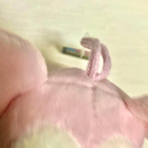 May include: A soft, plush toy featuring light pink fabric with a textured, fuzzy surface. The object has a small, curled, pink fabric loop attached to the top, and a small, rectangular tag is visible near the base of the loop.