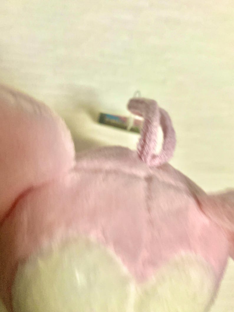 May include: A soft, plush toy featuring light pink fabric with a textured, fuzzy surface. The object has a small, curled, pink fabric loop attached to the top, and a small, rectangular tag is visible near the base of the loop.