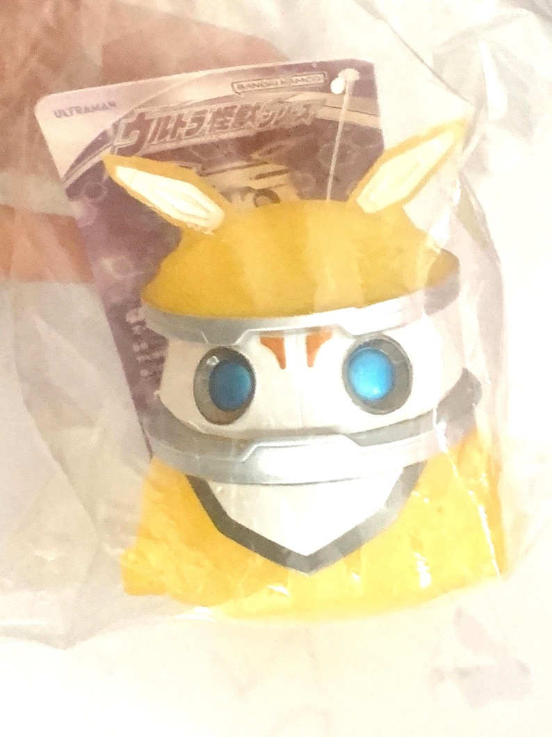 May include: A yellow and white Ultraman character figure with pointed ears and blue eyes, encased in a clear plastic bag with an attached cardboard tag featuring the text Ultraman and Japanese characters at the top of the packaging.
