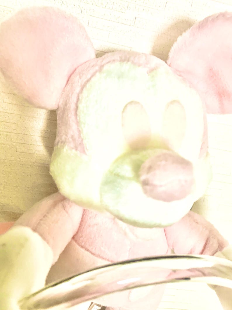 May include: A plush toy character with large rounded ears, a soft pink and pale green color palette, and a rounded snout. The stuffed figure features a smooth, fuzzy texture throughout its body, with prominent ears and a distinct facial structure.