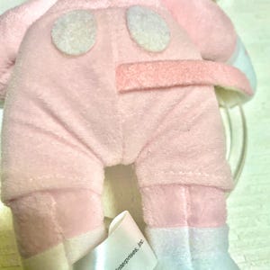 May include: A plush toy featuring a soft pink fabric body with two white circular patches on the back and a pink tail. A white fabric tag is attached to the lower portion, clearly displaying the printed text Disney Enterprises, Inc.