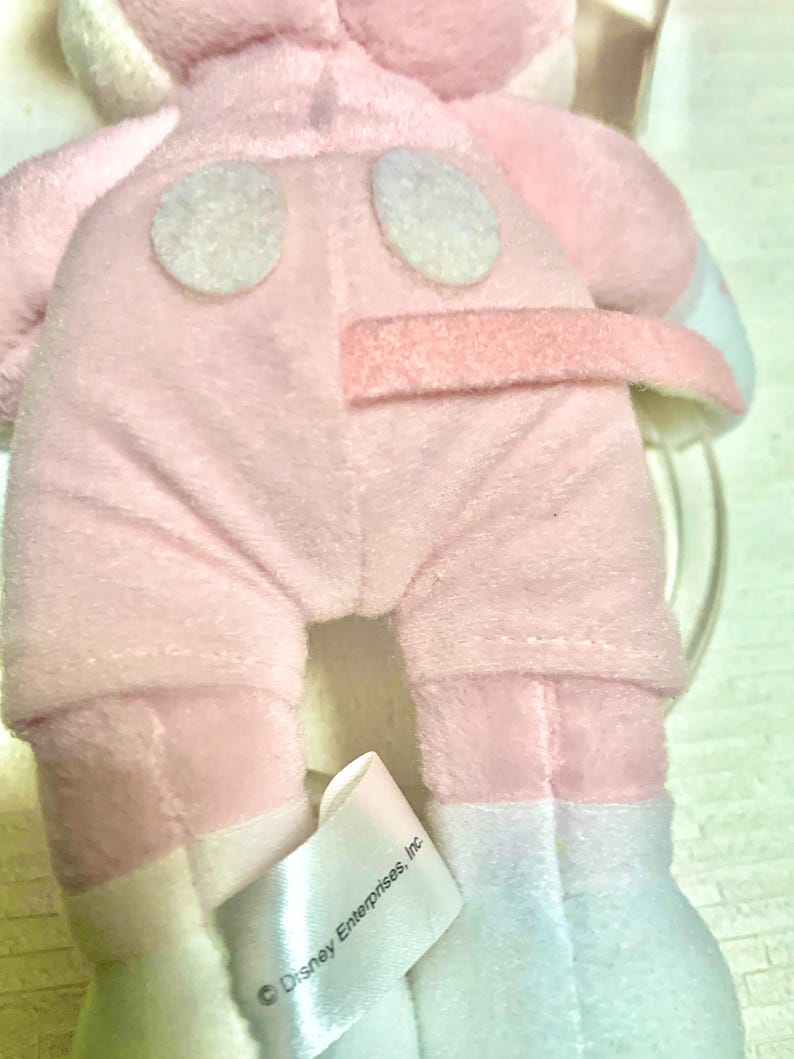 May include: A plush toy featuring a soft pink fabric body with two white circular patches on the back and a pink tail. A white fabric tag is attached to the lower portion, clearly displaying the printed text Disney Enterprises, Inc.