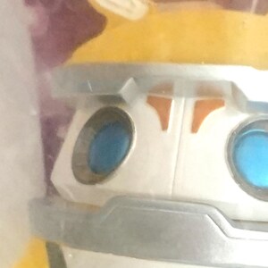 May include: A close-up of a stylized character figure featuring a white face with a silver brow and chin guard. The figure has two circular, bright blue eyes and a yellow top section with pointed ears. The design includes orange accents.
