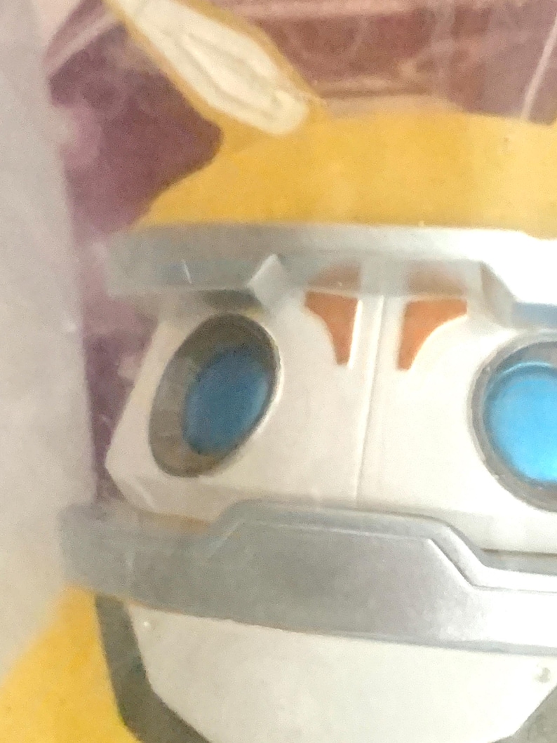 May include: A close-up of a stylized character figure featuring a white face with a silver brow and chin guard. The figure has two circular, bright blue eyes and a yellow top section with pointed ears. The design includes orange accents.