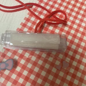 Pode incluir: Um porta-identifica&ccedil;&otilde;es de pl&aacute;stico transparente com um cord&atilde;o vermelho, sobre um fundo xadrez vermelho e branco. O suporte tem um fecho de velcro branco e um la&ccedil;o azul claro. O cord&atilde;o &eacute; vermelho vivo.