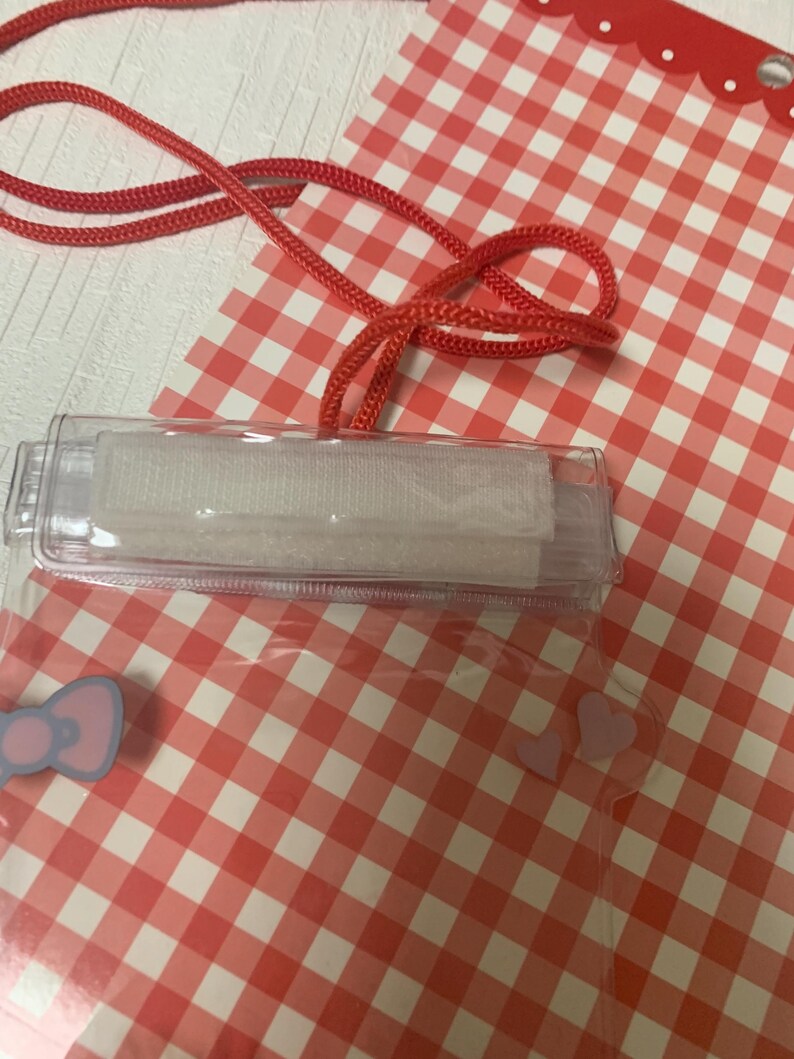 Pode incluir: Um porta-identifica&ccedil;&otilde;es de pl&aacute;stico transparente com um cord&atilde;o vermelho, sobre um fundo xadrez vermelho e branco. O suporte tem um fecho de velcro branco e um la&ccedil;o azul claro. O cord&atilde;o &eacute; vermelho vivo.