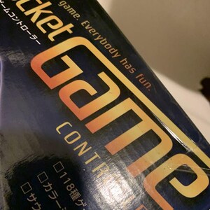 Pode incluir: Uma caixa de controlador de jogo azul marinho com as palavras "Pocket Game Controller" em letras brancas e douradas. A caixa tamb&eacute;m tem o texto "Let's play a game. Everybody has fun." em letras brancas.