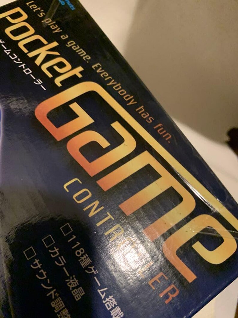 Pode incluir: Uma caixa de controlador de jogo azul marinho com as palavras "Pocket Game Controller" em letras brancas e douradas. A caixa tamb&eacute;m tem o texto "Let's play a game. Everybody has fun." em letras brancas.