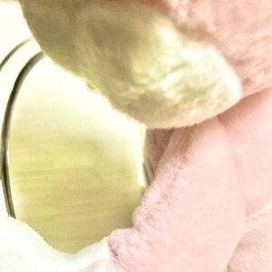 May include: A soft, plush toy featuring light pink and white fabric. The stuffed animal has a rounded, fuzzy texture with visible stitching along the arm and torso. A metallic, circular frame is positioned to the left of the plush figure.