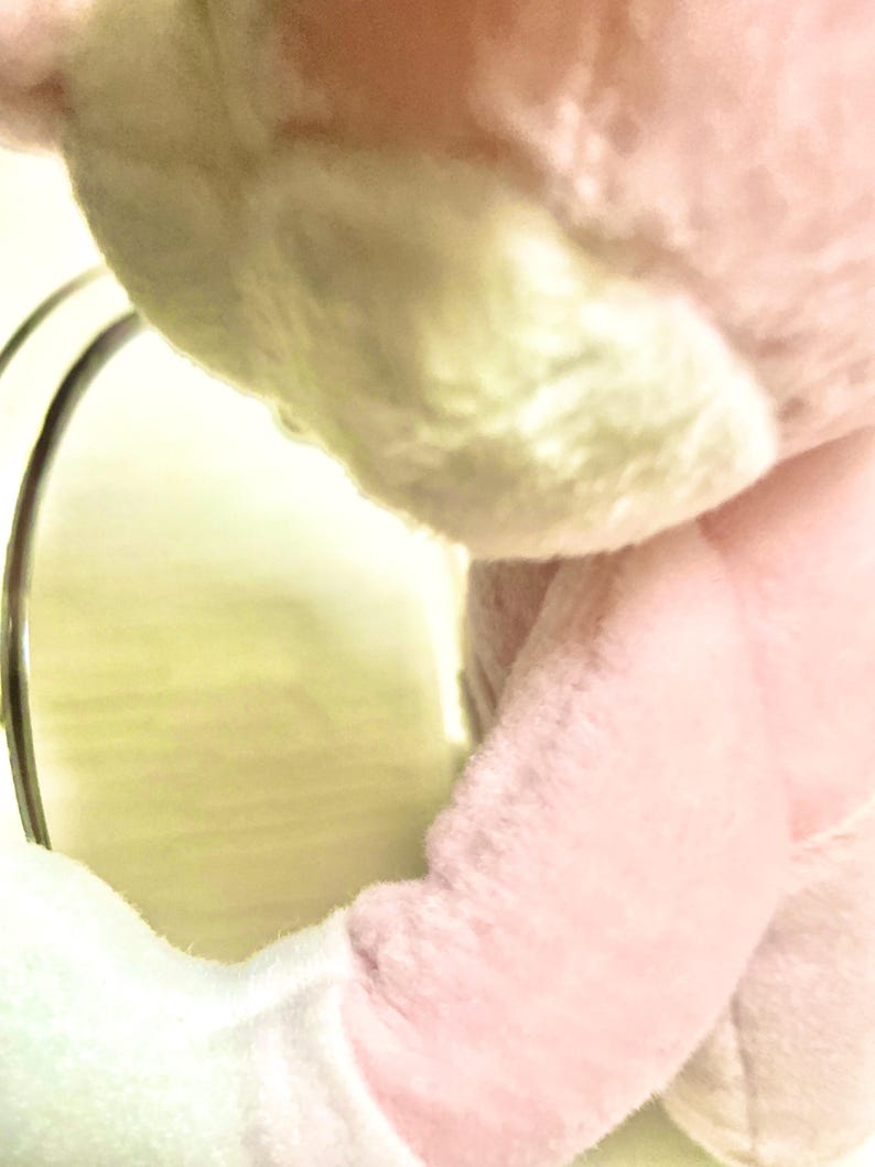 May include: A soft, plush toy featuring light pink and white fabric. The stuffed animal has a rounded, fuzzy texture with visible stitching along the arm and torso. A metallic, circular frame is positioned to the left of the plush figure.