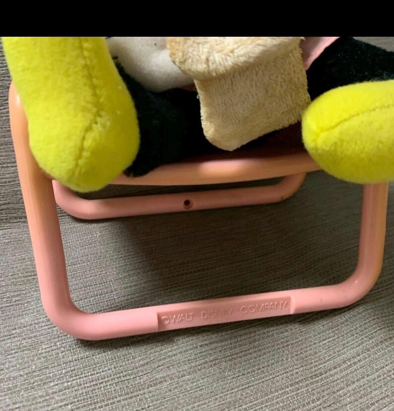 May include: A pink plastic chair with rounded corners, holding a plush toy with yellow, black, and beige elements. The text "WALT DISNEY COMPANY" is printed on the chair's frame. The toy's yellow hand and foot are visible.