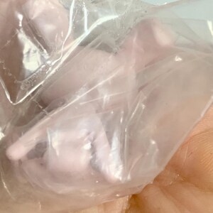 Puede incluir: Una peque&ntilde;a figura moldeada de color rosa p&aacute;lido encerrada en una bolsa de pl&aacute;stico transparente y arrugada. El objeto presenta contornos suaves y redondeados y una forma compacta y abstracta, visible a trav&eacute;s de la superficie transl&uacute;cida.