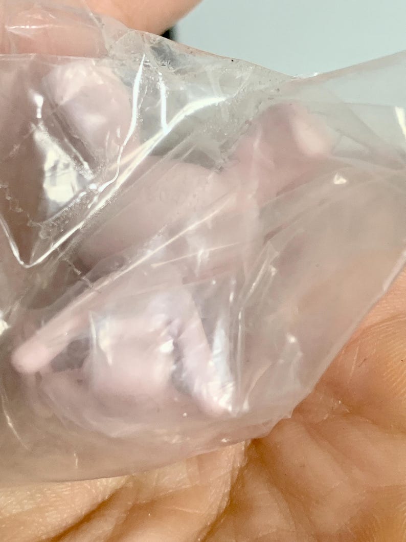 Puede incluir: Una peque&ntilde;a figura moldeada de color rosa p&aacute;lido encerrada en una bolsa de pl&aacute;stico transparente y arrugada. El objeto presenta contornos suaves y redondeados y una forma compacta y abstracta, visible a trav&eacute;s de la superficie transl&uacute;cida.