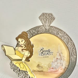 May include: A silver-toned metal pin shaped like a ring with a diamond top. The circular frame features embossed roses and depicts Belle reading a book, with a yellow background showing a castle and the text Beauty and the Beast Disney.