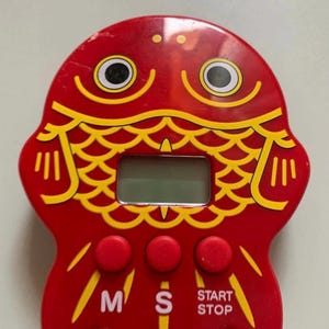 May include: A red fish-shaped kitchen timer with yellow scale details and black and white eyes. The timer has three red buttons and a small digital display. The buttons are labeled "M", "S", "START", and "STOP".