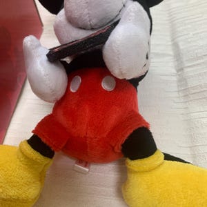 Puede incluir: Un peluche de Mickey Mouse con pantalones cortos rojos, zapatos amarillos y guantes blancos. El juguete tiene una cabeza en blanco y negro con grandes orejas negras. El juguete est&aacute; sobre una superficie blanca.