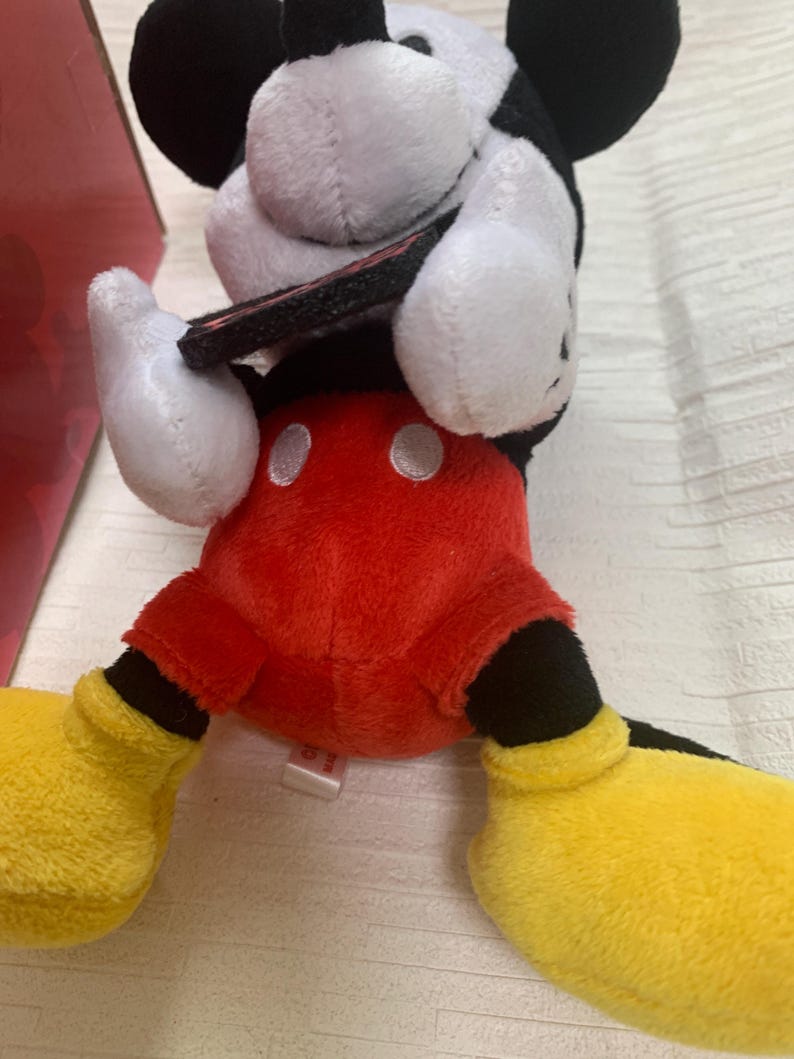 Puede incluir: Un peluche de Mickey Mouse con pantalones cortos rojos, zapatos amarillos y guantes blancos. El juguete tiene una cabeza en blanco y negro con grandes orejas negras. El juguete est&aacute; sobre una superficie blanca.