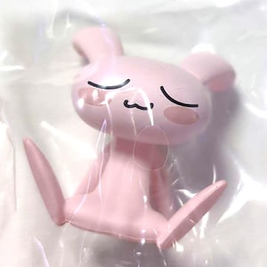 Puede incluir: Una peque&ntilde;a figura de pl&aacute;stico de color rosa claro que representa a una criatura sentada parecida a un conejo, con orejas largas, ojos cerrados y una boca sencilla. El personaje tiene mejillas redondas y est&aacute; envuelto en pl&aacute;stico transparente.