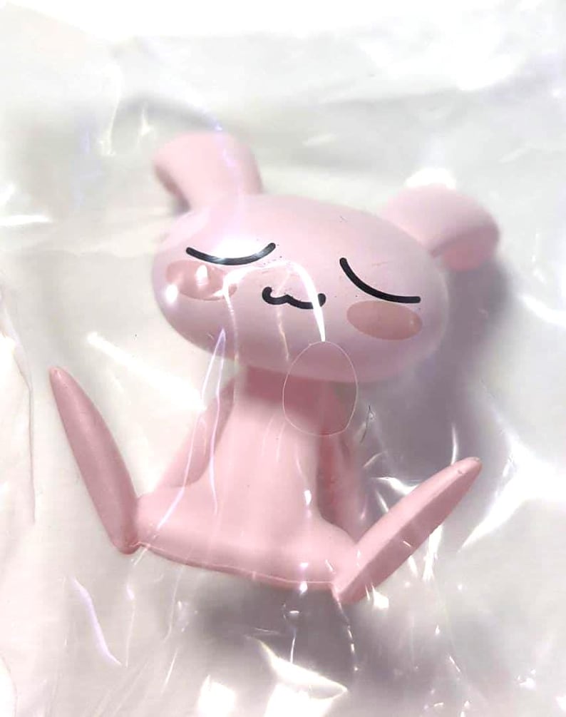 Puede incluir: Una peque&ntilde;a figura de pl&aacute;stico de color rosa claro que representa a una criatura sentada parecida a un conejo, con orejas largas, ojos cerrados y una boca sencilla. El personaje tiene mejillas redondas y est&aacute; envuelto en pl&aacute;stico transparente.