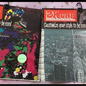 May include: Two 2XCAP headwear design packaging cards. The left card features a black background with colorful paint splatters and the text 'Customize your style to be cool'. The right card displays a black and white cityscape with the same text.