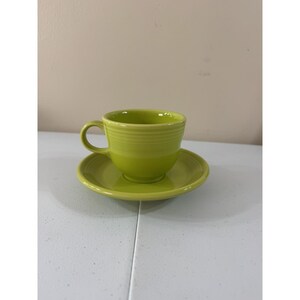 May include: A lime green ceramic teacup and matching saucer set. The teacup features a rounded handle and a series of horizontal ridges encircling the upper exterior. The saucer has a smooth, circular design with a slightly raised center rim.