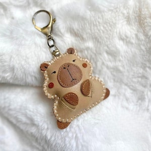 May include: A tan leather keychain shaped like a stylized bear with a darker brown snout, small ears, and stitched edges. The bear features a simple embroidered face and is attached to a bronze-colored metal lobster clasp for easy attachment.
