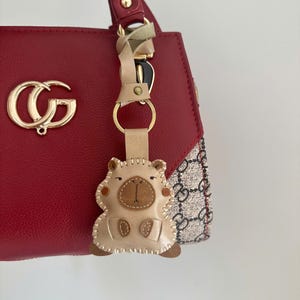 May include: A red handbag with a gold-toned logo emblem and a beige patterned side panel. A beige leather keychain shaped like a stylized animal with stitched details and a gold metal ring hangs from the red handle of the bag.