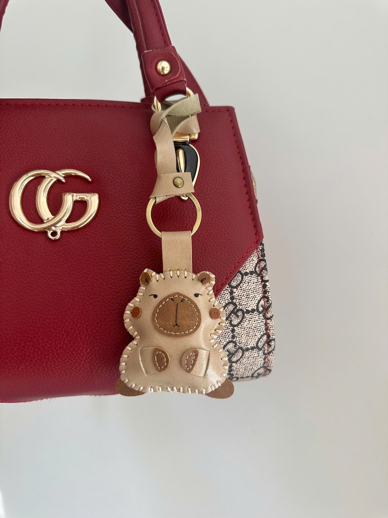May include: A red handbag with a gold-toned logo emblem and a beige patterned side panel. A beige leather keychain shaped like a stylized animal with stitched details and a gold metal ring hangs from the red handle of the bag.