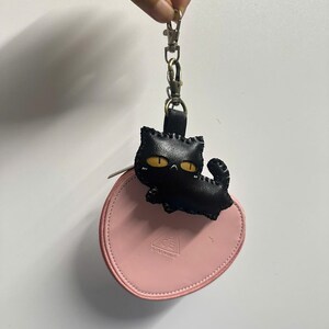 May include: A black leather cat keyring with yellow eyes is attached to a pink, round zipped pouch. The keyring is held by a silver clasp. The pouch has the text "CE" printed on it.