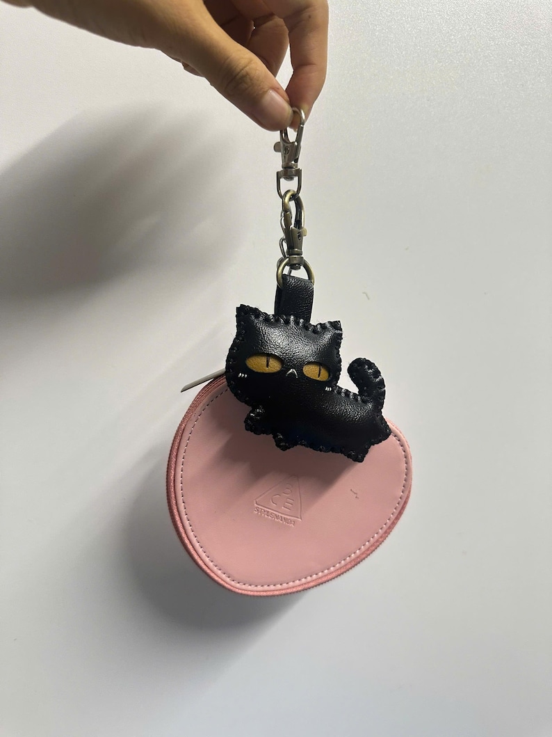 May include: A black leather cat keyring with yellow eyes is attached to a pink, round zipped pouch. The keyring is held by a silver clasp. The pouch has the text "CE" printed on it.