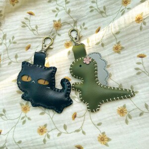 May include: Two leather keyrings: a black cat with yellow eyes and a green dinosaur with a grey back. Both have metal clasps and are stitched with white thread. They rest on cream fabric with a floral pattern.