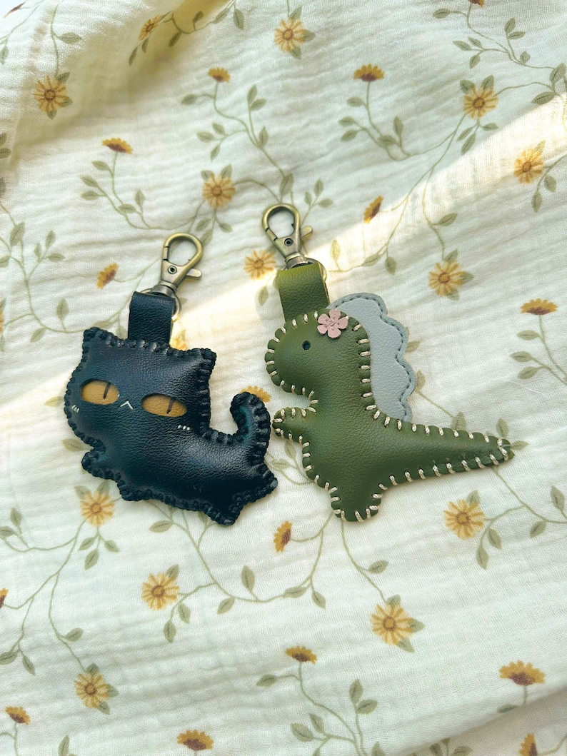 May include: Two leather keyrings: a black cat with yellow eyes and a green dinosaur with a grey back. Both have metal clasps and are stitched with white thread. They rest on cream fabric with a floral pattern.