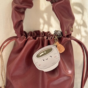 May include: A burgundy leather drawstring bag with a gathered top and a looped handle. A small, grey and white charm with a smiling face and a small, tan charm hang from the handle.