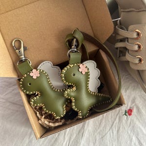 May include: Two olive green leather dinosaur keyrings with pink flower accents and white wing details. The keyrings are in a cardboard box with a pair of shoes in the background. The keyrings have gold stitching.
