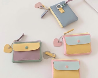 Può includere: Quattro piccole pochette rettangolari in colori pastello. Una pochette blu ha un cinturino da polso. Tre pochette presentano una patta con bottone a pressione, una tasca gialla o blu e un ciondolo a forma di cuore su una catenella.