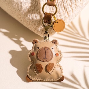 May include: A beige leather keychain shaped like a bear with a dark brown snout, small ears, and stitched details. It features a bronze-colored metal lobster clasp, a circular leather tag, and a brown leather strap attached to a metal ring.