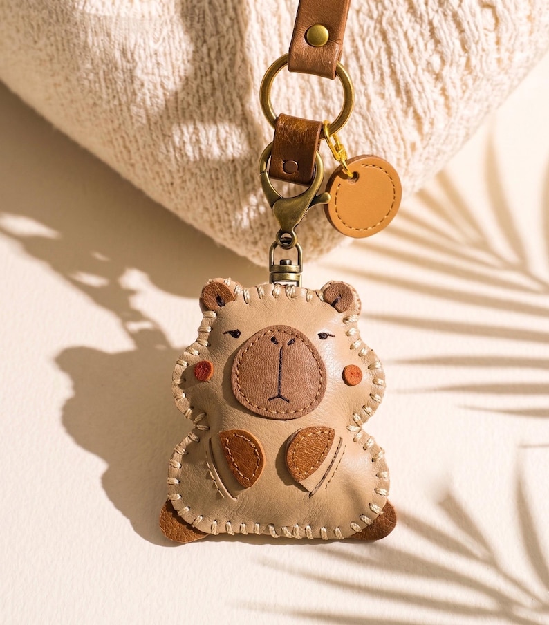 May include: A beige leather keychain shaped like a bear with a dark brown snout, small ears, and stitched details. It features a bronze-colored metal lobster clasp, a circular leather tag, and a brown leather strap attached to a metal ring.