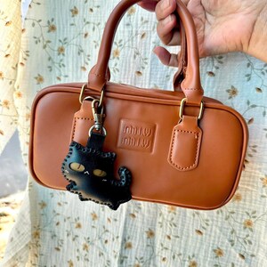 May include: A tan rectangular handbag with a top handle and gold-coloured hardware. The bag has a black cat-shaped keychain. The brand name is embossed on the front of the bag.