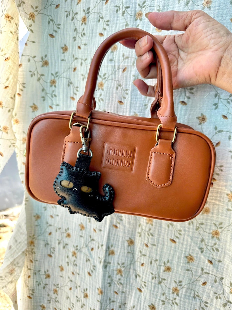 May include: A tan rectangular handbag with a top handle and gold-coloured hardware. The bag has a black cat-shaped keychain. The brand name is embossed on the front of the bag.
