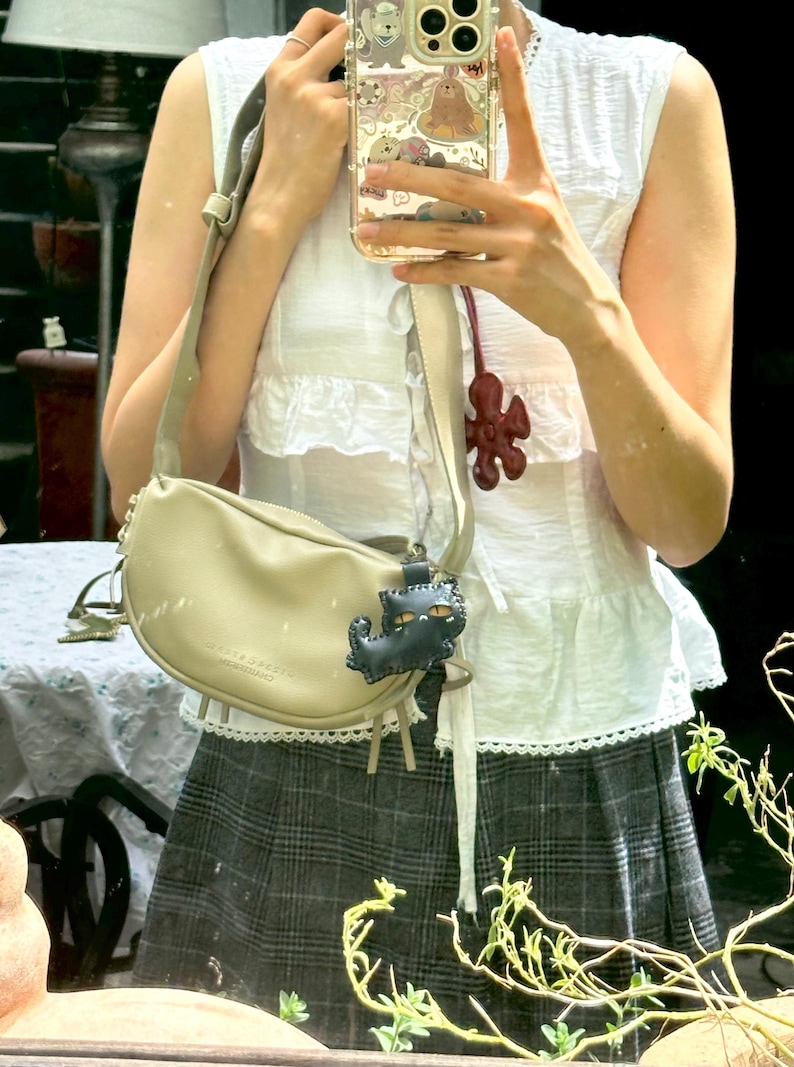 May include: A beige crescent-shaped shoulder bag with a matching strap. The bag has a small, dark blue cat-shaped charm and a zip closure. The bag is worn over a white top and a plaid skirt.