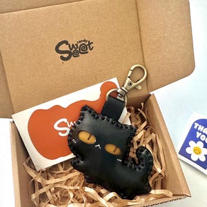 May include: A black leather cat keyring with yellow eyes and a stitched outline, presented in a cardboard box with the "Sweet Candy" logo. The box includes an orange card and a "Thank You" sticker.