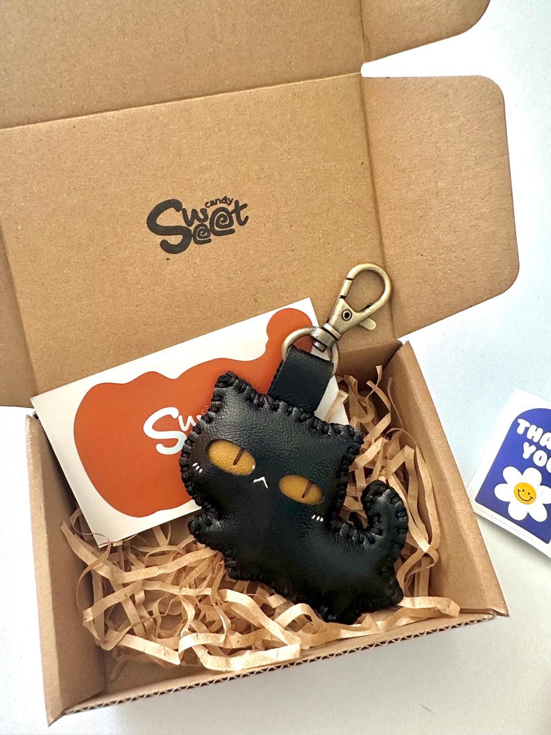 May include: A black leather cat keyring with yellow eyes and a stitched outline, presented in a cardboard box with the "Sweet Candy" logo. The box includes an orange card and a "Thank You" sticker.