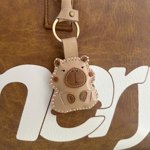 May include: A light tan leather keychain shaped like a capybara with stitched edges, a dark brown nose, and small circular cheeks. The keychain is attached to a gold-colored metal ring and a leather strap, hanging against a brown bag with white text.