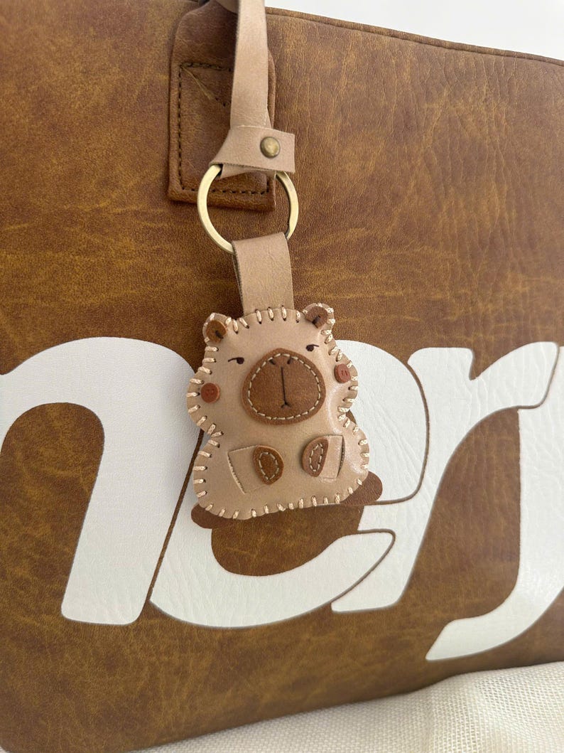 May include: A light tan leather keychain shaped like a capybara with stitched edges, a dark brown nose, and small circular cheeks. The keychain is attached to a gold-colored metal ring and a leather strap, hanging against a brown bag with white text.